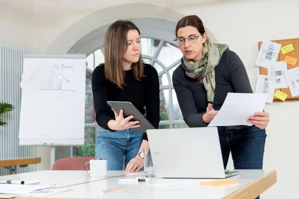 Zwei Menschen blicken auf einen Laptop und besprechen etwas. Eine hat ein Tablet in der Hand, die andere ein Blatt Papier.