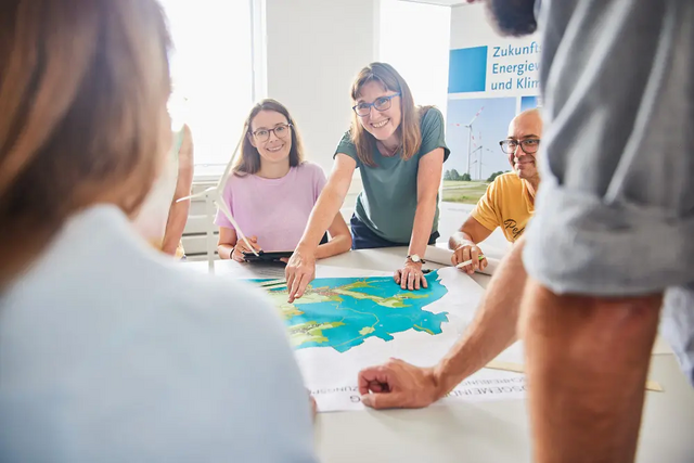 Mehrere Menschen sitzen an einem Tisch. Auf dem Tisch liegt ein großer Plan. Darauf steht ein Windrand. Jemand deutet mit dem Finger auf den Plan.