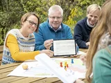 Vier Menschen sitzen an einem Tisch. Zwei davon zeigen auf ein Tablet und erklären etwas.