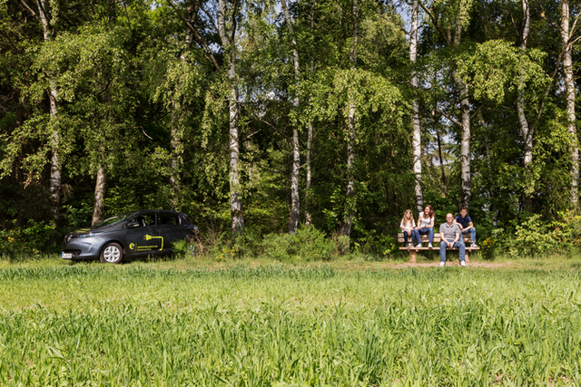 Das Foto zeigt ein E-Auto und eine Familie im Grünen