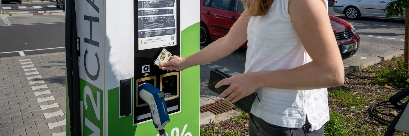 An einer Schnellladestation für Elektrofahrzeuge auf einem Supermarkt-Parkplatz hält ein Mensch eine Debitkarte an die Säule