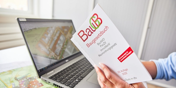Auf dem Tisch liegt ein Plan, darauf steht ein Laptop. Eine Person sitzt vor dem Laptop und hält das Baugesetzbuch in den Händen.