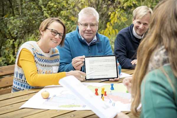 Vier Menschen sitzen an einem Tisch. Zwei davon zeigen auf ein Tablet und erklären etwas.