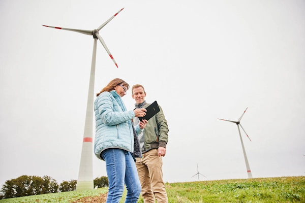 Zwei Menschen stehen auf einem Feld. Im Hintergrund stehen zwei Windräder. Die beiden Menschen blicken auf ein tablet und besprechen sich.