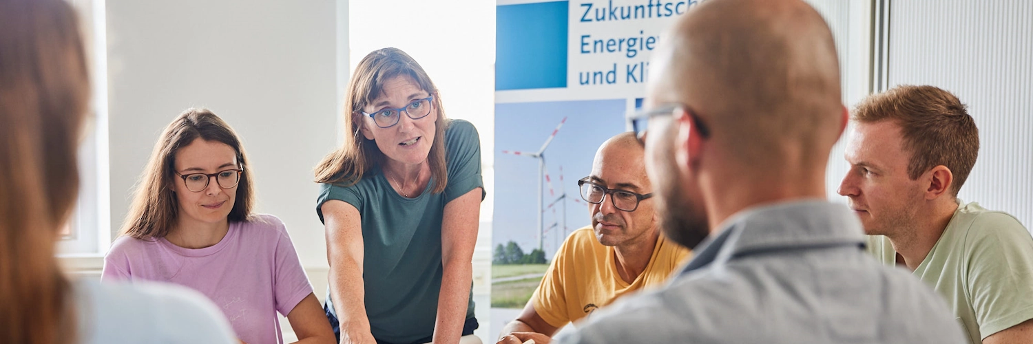 Mehrere Menschen sitzen an einem Tisch. Eine Person zeigt auf einen Plan, der auf dem Tisch liegt. Die anderen schauen die Person an oder auf den Plan.