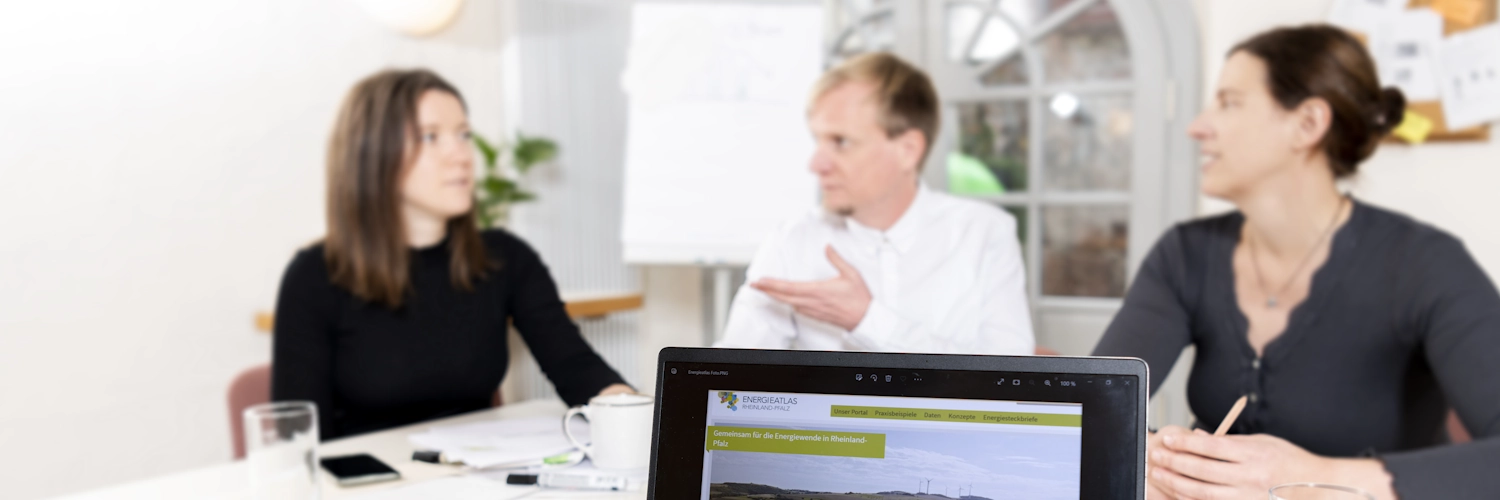 Drei Menschen sitzen an einem Tisch und diskutieren, sie sind im Hintergrund zu sehen. Im Vordergrund ist ein Laptop zu sehen, die Website des Energieatlas RLP ist geöffnet.