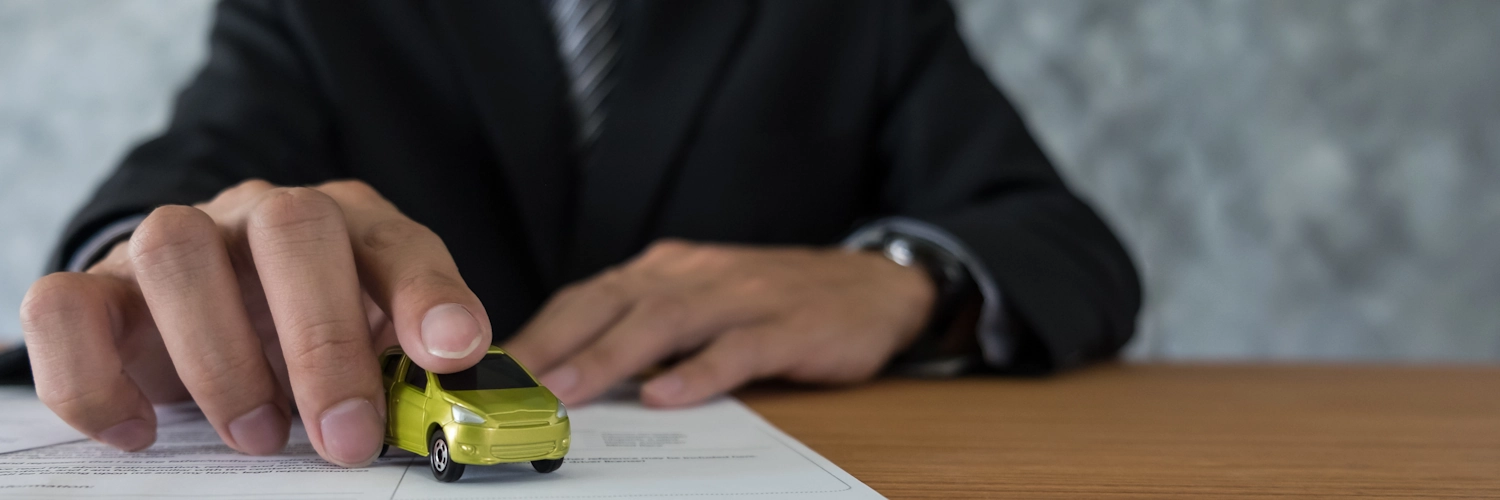 Eine Person mit Jacket sitzt an einem Tisch. Auf dem Tisch liegt ein Blatt Papier, darauf steht ein Spielzeug-Auto. Die Person hält das Auto mit dem Zeigefinger fest bzw. bewegt es.