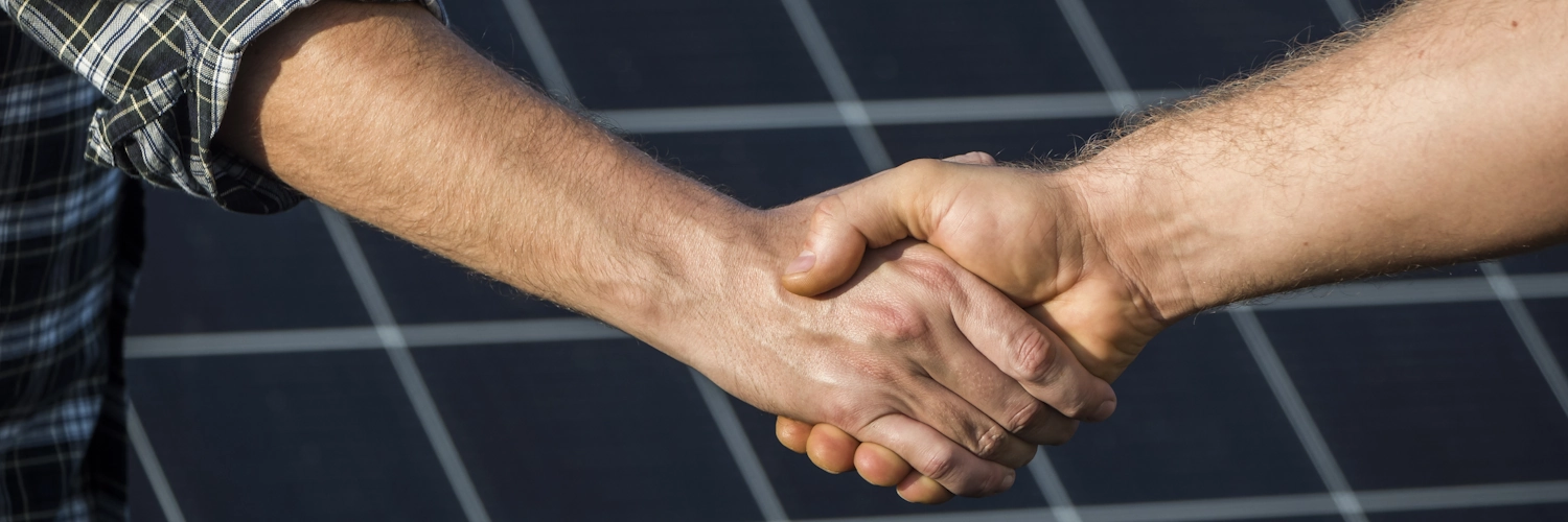 Zwei Menschen geben sich die Hand in Form eines Handschlags. Im Hintergrund sind Solarpanele zu sehen.