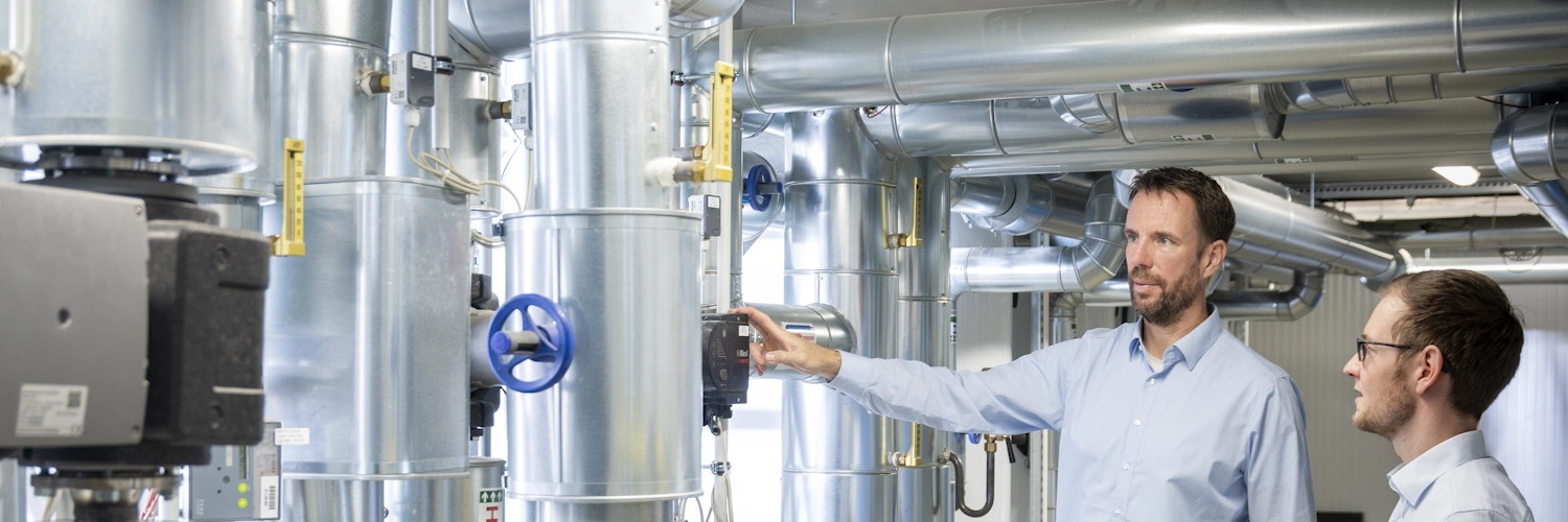 Zwei Menschen stehen im Heizraum eines kommunalen Gebäudes und lesen Daten ab.